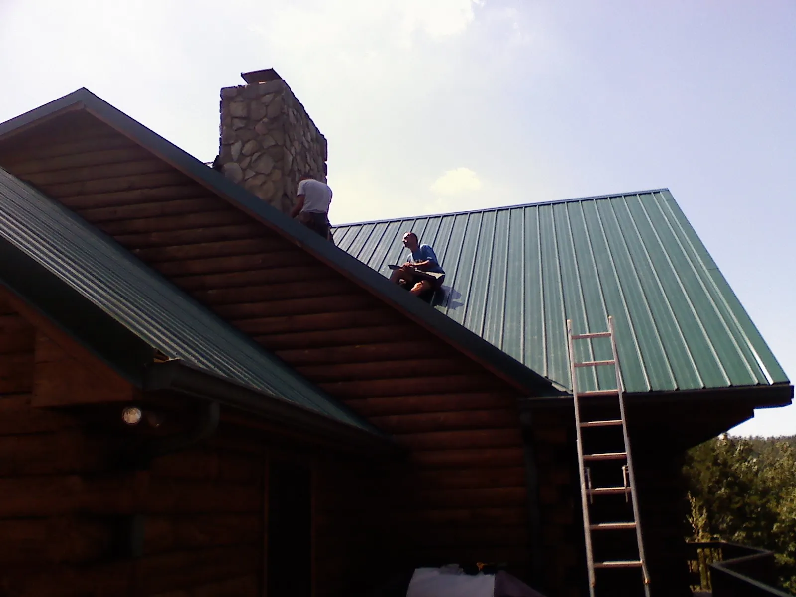 Roofers installing green metal panels beside a stone chimney on a log home in Lindon